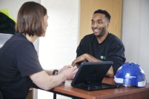 A Black man smiling while talking to a white person across a desk with a laptop and a blue hard hat.