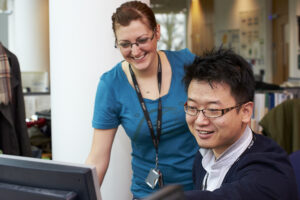 A man and a woman in an office setting, both smiling and looking at a computer monitor.