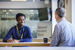 Two office scenes, one showing a Black man smiling while talking to a white man across a desk in front of a National Incident Room