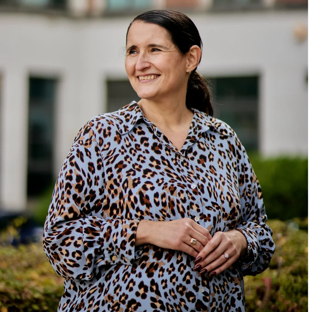 A smiling woman wearing a light blue leopard-print shirt, standing outdoors and looking to the left.