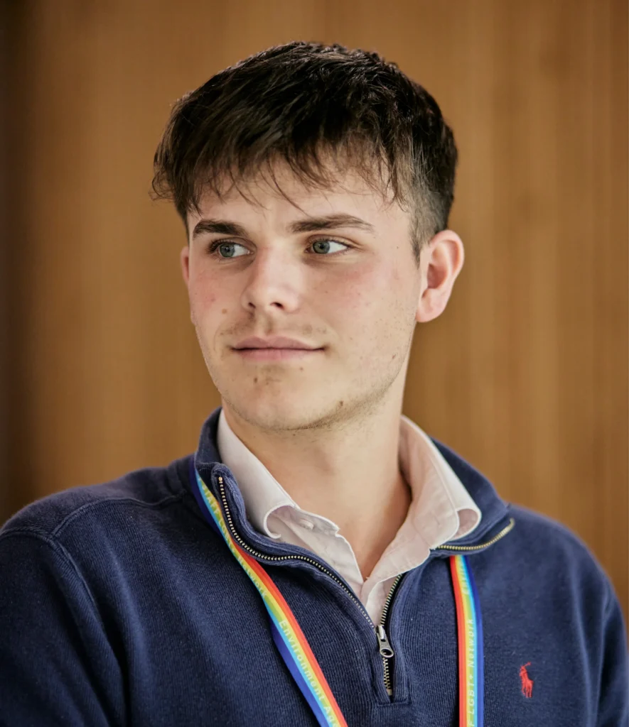A young man wearing a navy blue quarter-zip sweater and a rainbow lanyard over a white shirt, looking to the left.